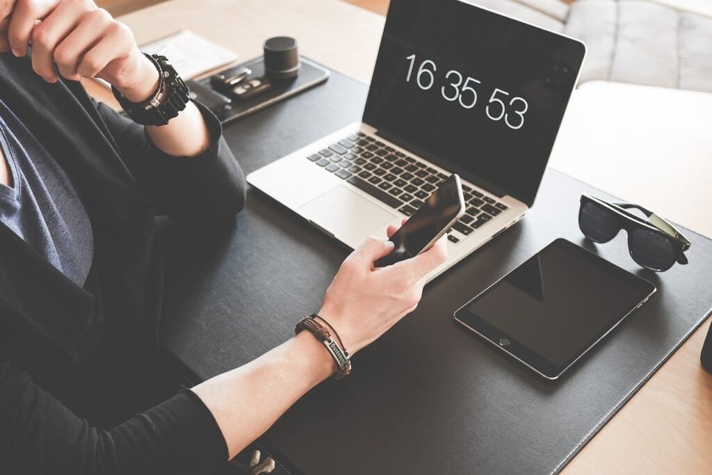 Person sitting at a desk holding an iPhone with a mac book and iPad on the desk.