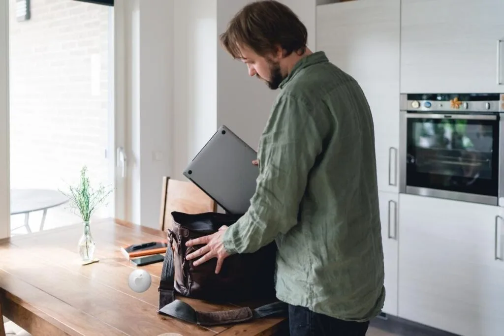 Ways to use an AirTag for your laptop bag A man in his kitchen putting a laptop in a bag with an AirTag on the table as an example to use an AirTag with a laptop bag.