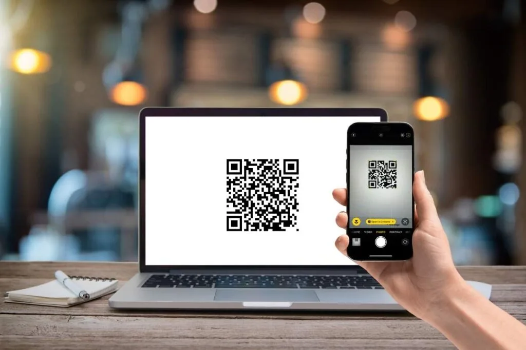 A laptop screen with a QR code on the screen and a person holding an iPhone scanning the QR code as an example of how do you scan a QR code on iPhone.