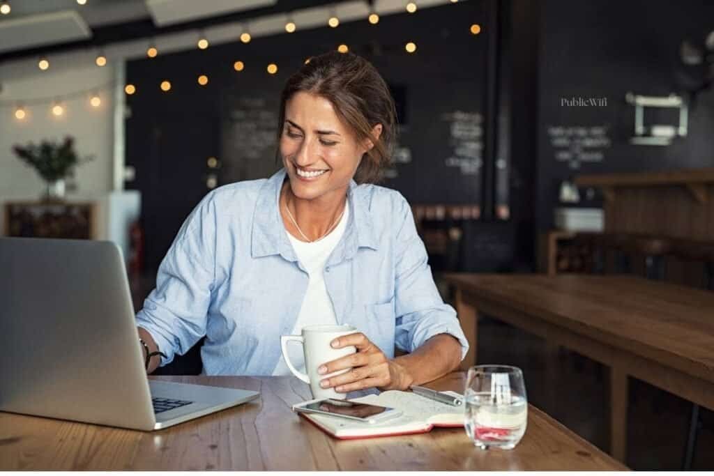 Woman sitting at a coffee shop working on a laptop with her iPhone next to her as an example of using personal hotspot instead of public wifi